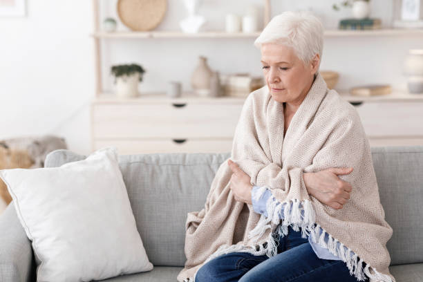 Senior woman feeling cold, wrapped tightly in a cozy blanket on the sofa.