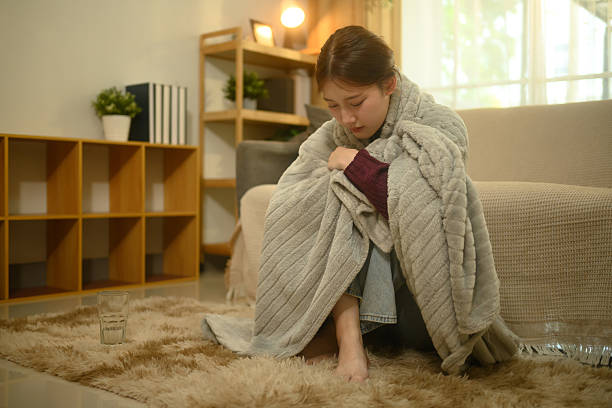Young woman huddling sadly in a cozy grey blanket on the floor.