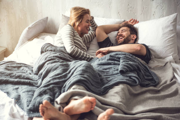 Happy couple laughing in bed, cozy under a soft grey blanket.