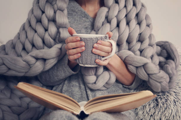 Person cozy in a chunky knit blanket, reading a book with a hot drink.