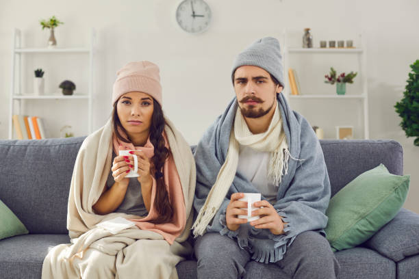Cold couple on sofa, wrapped in warm blankets, drinking hot beverages.