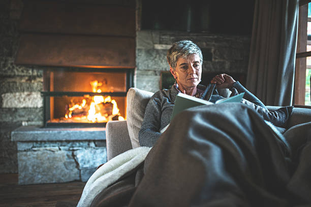 Woman reading a book by the fireplace, warm and cozy under a blanket.