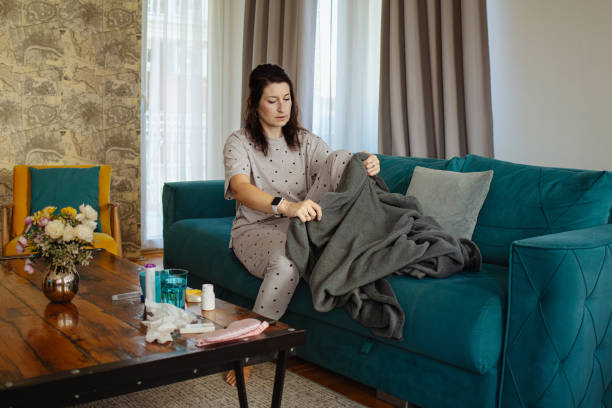 Woman in pajamas folding a soft grey blanket on a teal couch at home.
