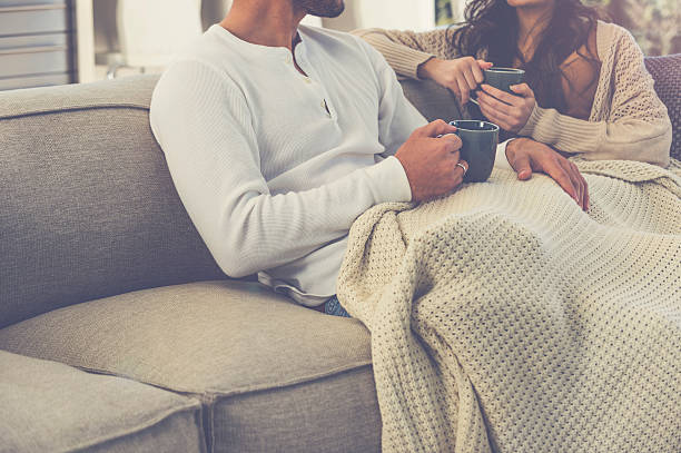 Couple relaxing on couch with a cozy knitted blanket, enjoying warm drinks.