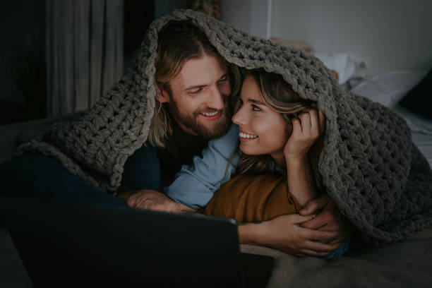 Happy couple snuggling under a chunky knitted blanket while watching laptop together.