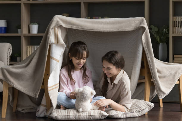 Two happy girls playing with a teddy bear inside a cozy blanket fort.