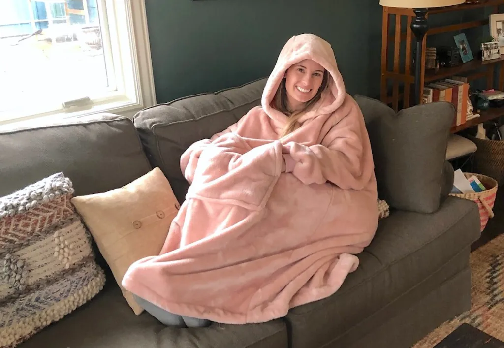 Happy woman in a comfy pink hooded wearable blanket relaxing on a couch.