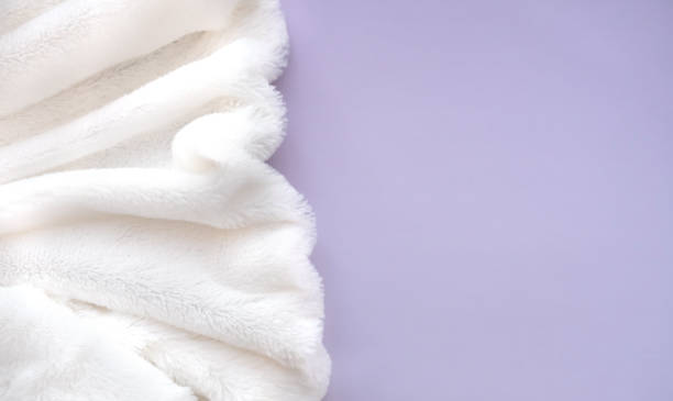 Soft white fluffy blanket draped against a calming purple background. Cozy blanket texture.