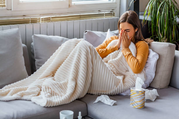 Sick woman comfortably resting on sofa, covered by a plush blanket, blowing her nose.