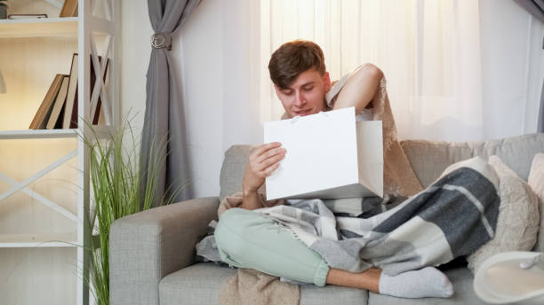 Man relaxing on sofa, cozy under a plaid blanket, opening a gift bag.