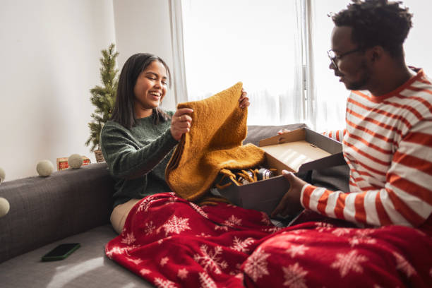 Couple opening gifts, cozy under a red snowflake blanket on the couch.